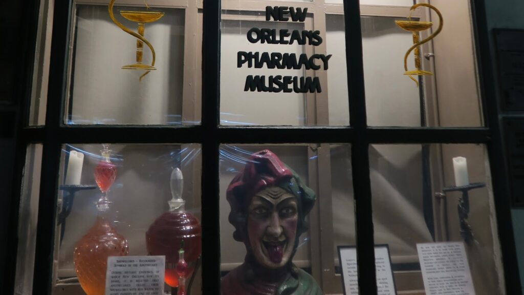 Window display at the New Orleans Pharmacy Museum featuring colorful glass bottles, candles, and a carved figure with exaggerated facial features and tongue sticking out. The museum name is painted on the glass above the display.