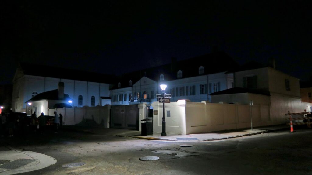 The Old Ursuline Convent in New Orleans at night, with its white walls, shuttered windows, and cross-topped roof dimly lit by a streetlamp on the corner. Shadows and darkened streets give the historic building a mysterious, eerie atmosphere.