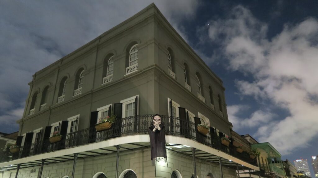 The LaLaurie Mansion in New Orleans’ French Quarter, photographed at night under a cloudy sky. The historic gray building has black shutters, wrought-iron balconies with flower boxes, and a spooky figure hanging from the railing.