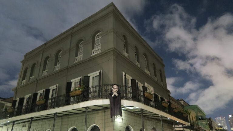 The LaLaurie Mansion in New Orleans’ French Quarter, photographed at night under a cloudy sky. The historic gray building has black shutters, wrought-iron balconies with flower boxes, and a spooky figure hanging from the railing.