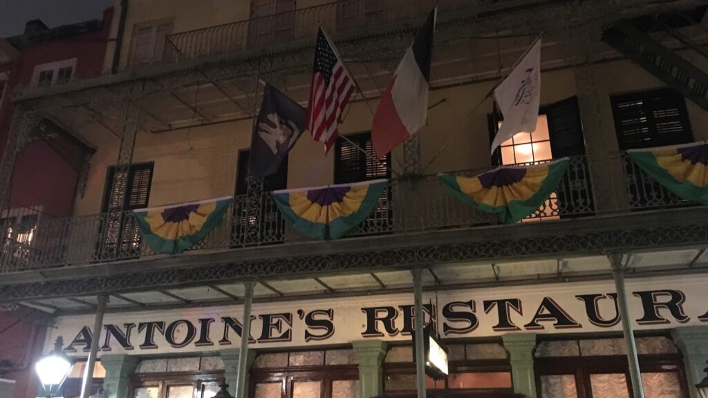 Exterior of Antoine’s Restaurant in New Orleans at night, decorated with Mardi Gras bunting and flags above its wrought-iron balcony.