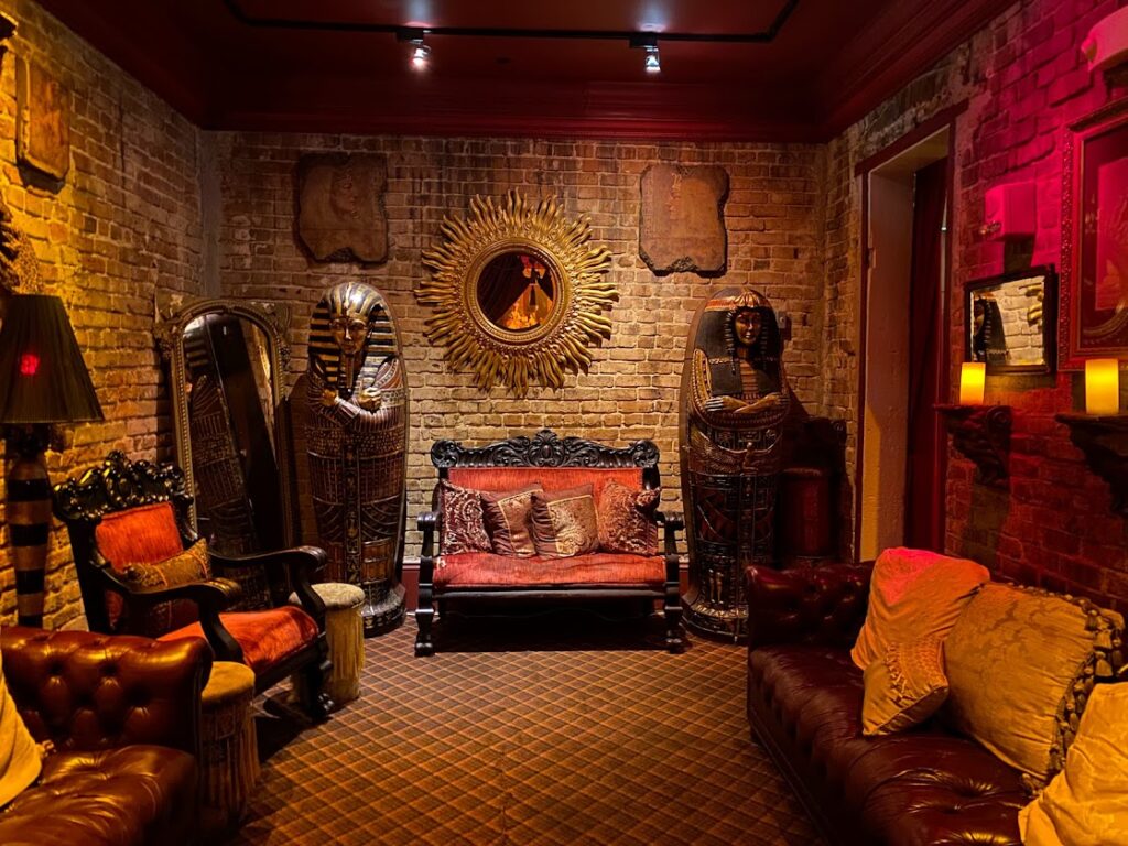 A dimly lit, Egyptian-themed lounge with brick walls, ornate vintage furniture, sarcophagus statues on either side of a red velvet settee, and warm candlelight creating a mysterious, spooky atmosphere in the upstairs area of Muriel's.