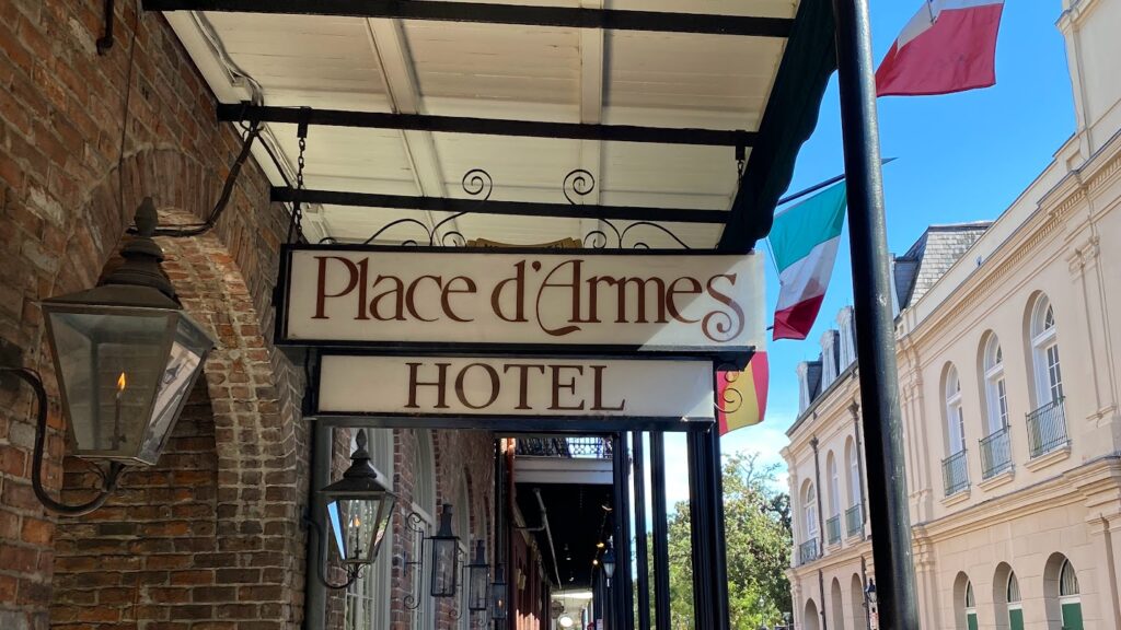 Hanging sign for the Place d’Armes Hotel in New Orleans’ French Quarter, with brick walls, wrought iron lanterns, and colorful flags along the street on a sunny day.