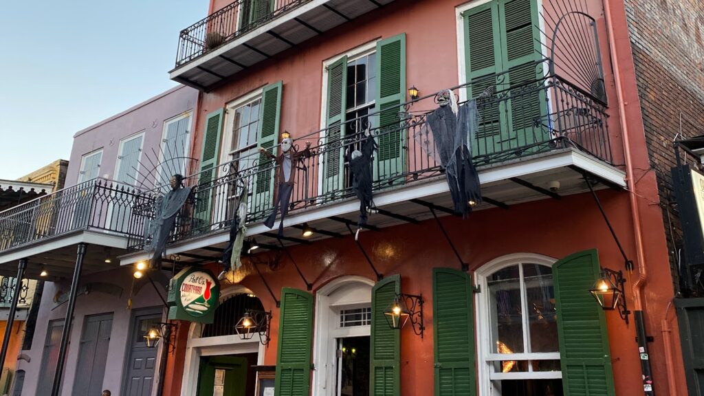 Exterior of Pat O’Brien’s Courtyard Restaurant in New Orleans’ French Quarter, decorated with spooky hanging skeleton figures and ghouls on the balcony railing, with green shutters and wrought-iron balconies.