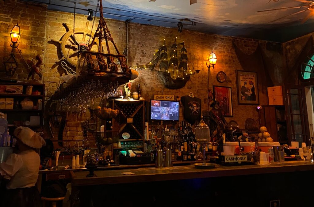 Interior of Pirate’s Alley Café in New Orleans, featuring a pirate-themed bar with a ship-shaped chandelier, hanging wine glasses, a model ship on the wall, bottles of liquor, and a sign that reads “Cash Only.” Warm lighting and nautical décor create a spooky, old-world atmosphere.