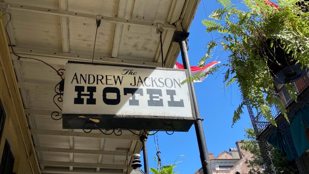 Hanging sign for the Andrew Jackson Hotel in New Orleans, mounted under a covered balcony with plants and a clear blue sky in the background.