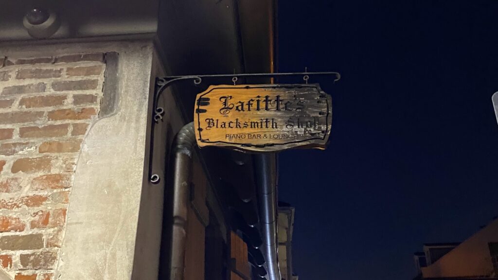 Weathered wooden sign for Lafitte’s Blacksmith Shop Bar in New Orleans, hanging from a wrought iron bracket against a brick wall at night.