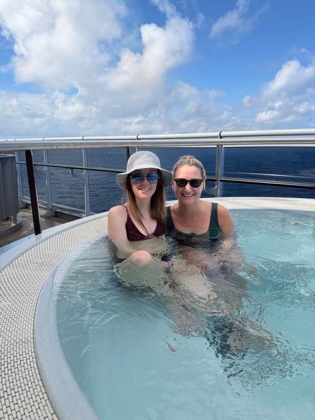 Two friends enjoying a relaxing dip in a hot tub aboard Virgin Voyages’ Scarlet Lady cruise ship.