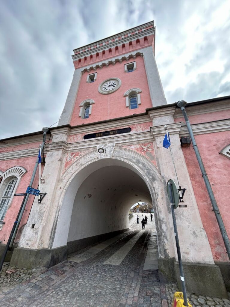 Pink historic gatehouse and clock tower in Suomenlinna, Finland, with a large arched passageway leading through the building and flags displayed on either side.