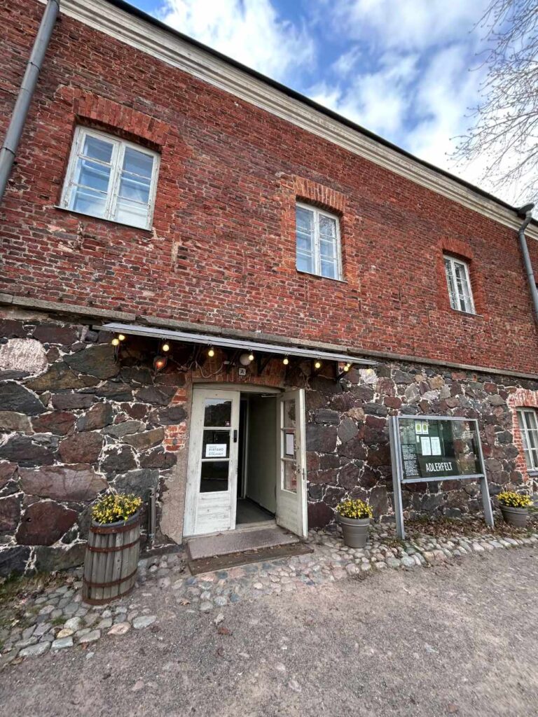 Historic two-story brick and stone building in Suomenlinna, Finland, with a white entrance door, small windows, and a cobblestone walkway in front.
