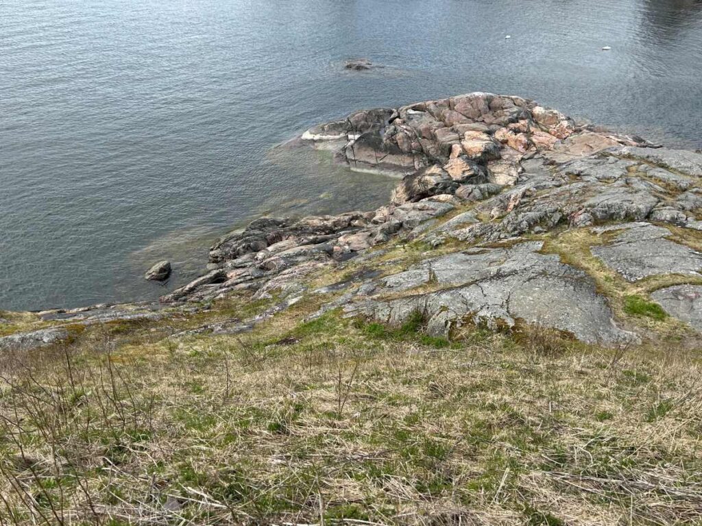 Rocky shoreline overlooking calm water in Suomenlinna, Finland, with weathered stone outcrops and sparse grass along the coast.