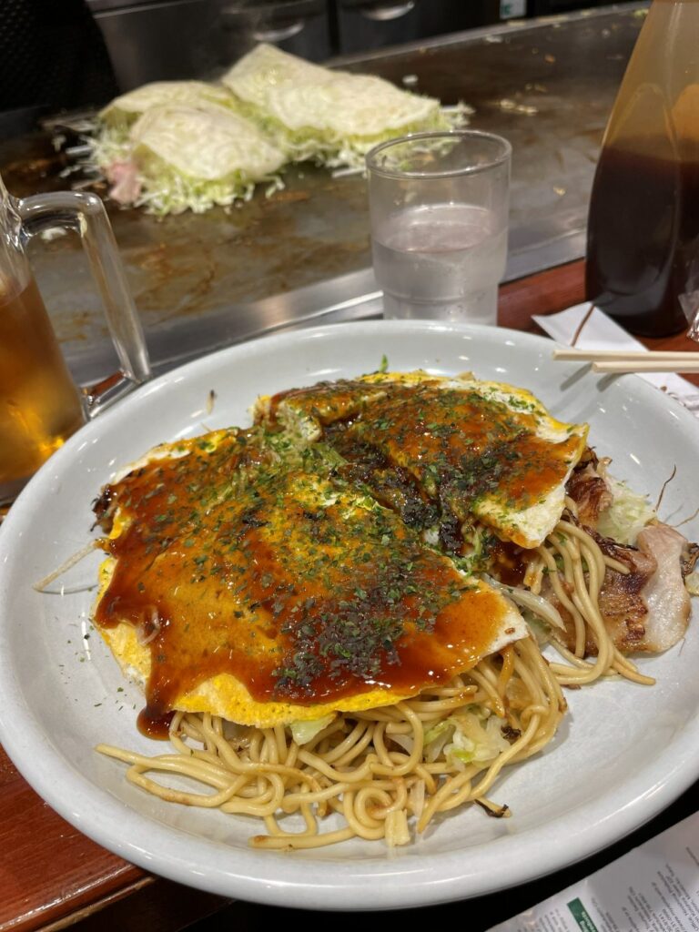 Traditional Hiroshima-style okonomiyaki layered with noodles, cabbage, egg, meat, and savory toppings.
